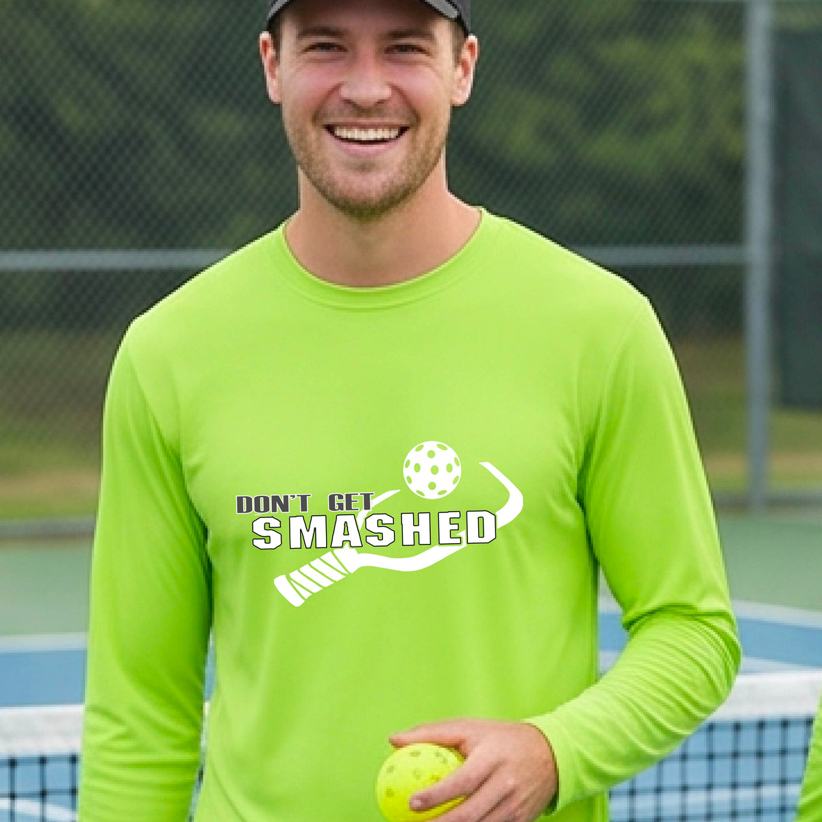 A man wearing a lime green long-sleeve t-shirt with a white graphic with the text 'DON'T GET SMASHED' and a white pickleball paddle and colorful pickleall graphic. 