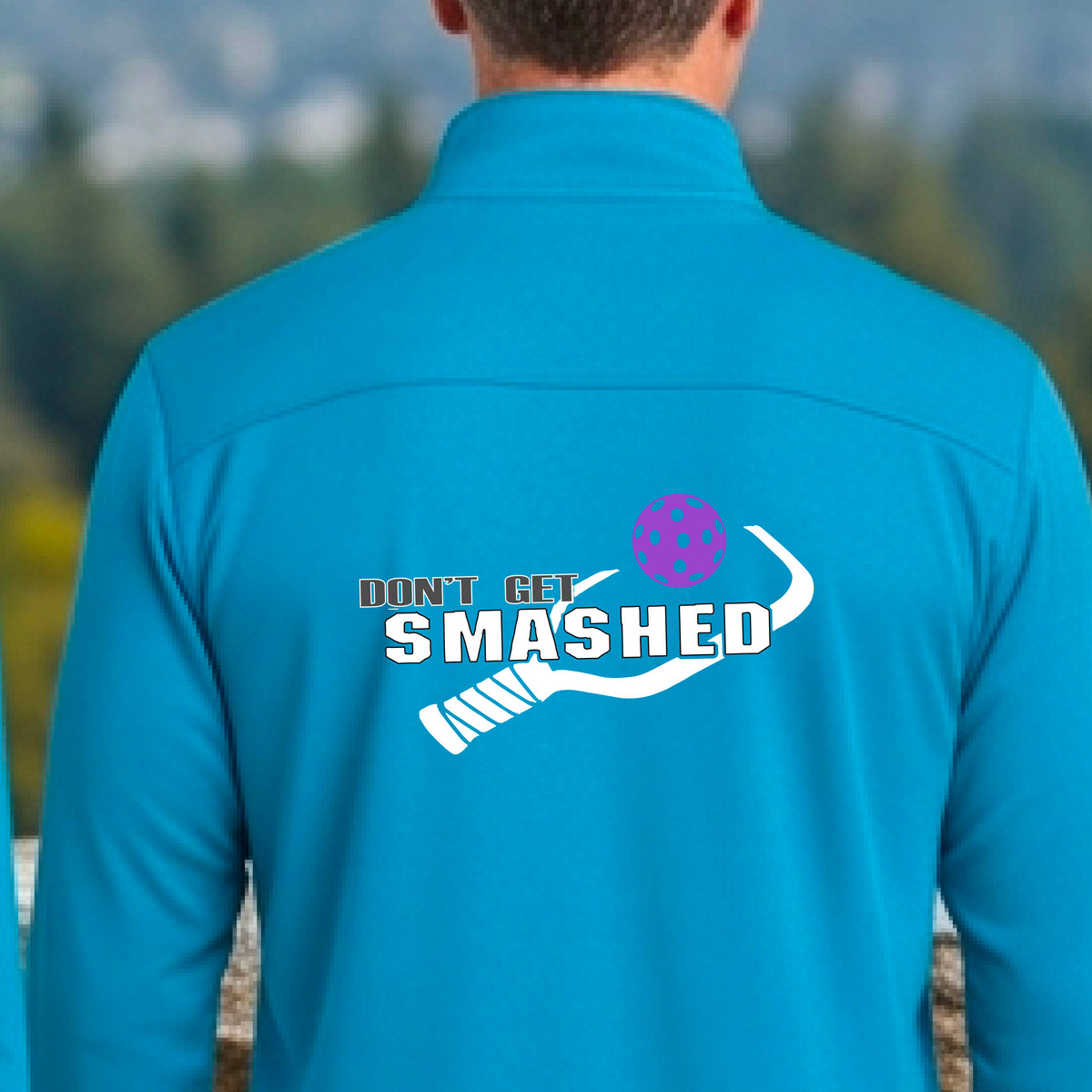 A man viewed from behind wearing a bright blue athletic quarter-zip jacket on a pickleball court, featuring a white graphic of a text 'DON'T GET SMASHED' and a white pickleball graphic and a colorful pickleball. 