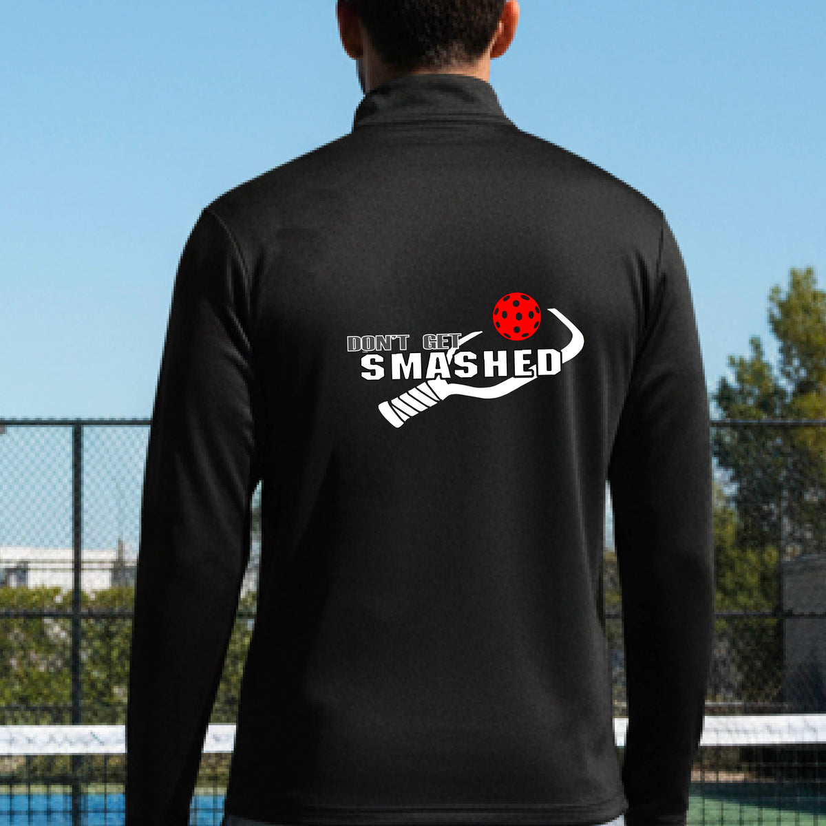 A man viewed from behind wearing a black athletic quarter-zip jacket on a pickleball court, featuring a white graphic of a text 'DON'T GET SMASHED' and a white pickleball graphic and a colorful pickleball. 