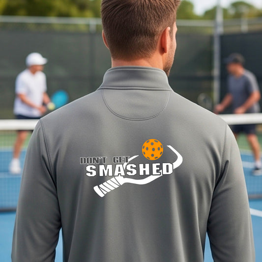 A man viewed from behind wearing a gray athletic quarter-zip jacket on a pickleball court, featuring a white graphic of a text 'DON'T GET SMASHED' and a white pickleball graphic and a colorful pickleball. 