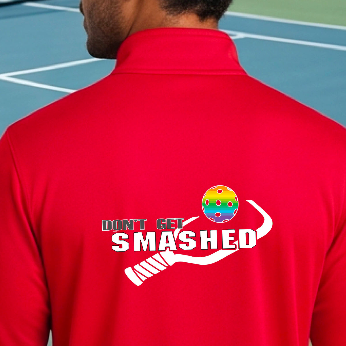 A man viewed from behind wearing a bright red athletic quarter-zip jacket on a pickleball court, featuring a white graphic of a text 'DON'T GET SMASHED' and a white pickleball graphic and a colorful pickleball. 