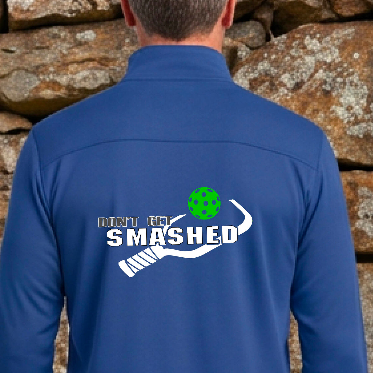 A man viewed from behind wearing a bright royal lbue athletic quarter-zip jacket  featuring a white graphic of a text 'DON'T GET SMASHED' and a white pickleball graphic and a colorful pickleball. 