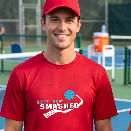 A smiling man wearing a red t-shirt on a pickleball court. The t-shirt features a graphic with a colorful pickleball and the white text 'DON'T GET SMASHED' with a pickleball paddle icon.