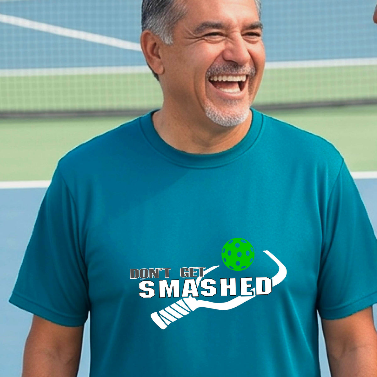 A smiling man wearing a teal t-shirt on a pickleball court. The t-shirt features a graphic with a colorful pickleball and the white text 'DON'T GET SMASHED' with a pickleball paddle icon.