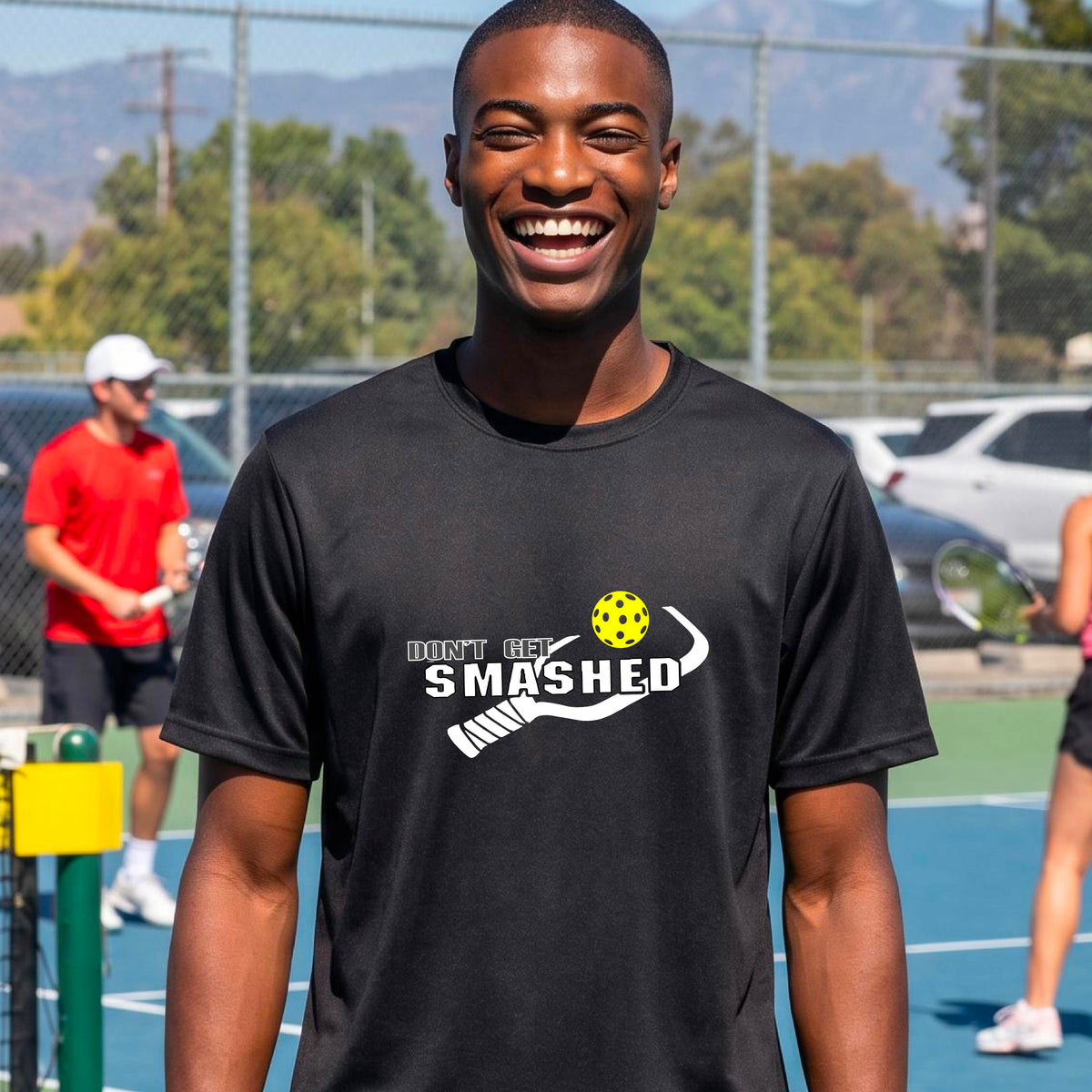A smiling man wearing a black t-shirt on a pickleball court. The t-shirt features a graphic with a colorful pickleball and the white text 'DON'T GET SMASHED' with a pickleball paddle icon.