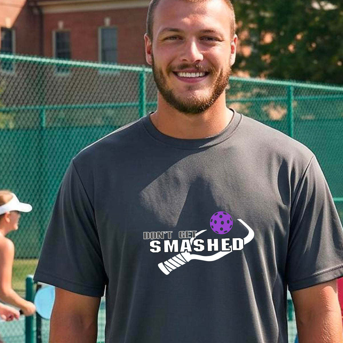 A smiling man wearing a dark gray t-shirt on a pickleball court. The t-shirt features a graphic with a colorful pickleball and the white text 'DON'T GET SMASHED' with a pickleball paddle icon.
