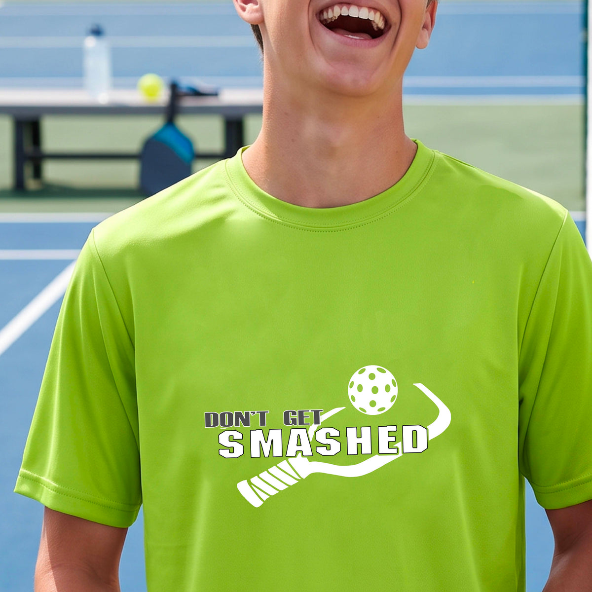 A smiling man wearing a lime green t-shirt on a pickleball court. The t-shirt features a graphic with a colorful pickleball and the white text 'DON'T GET SMASHED' with a pickleball paddle icon.