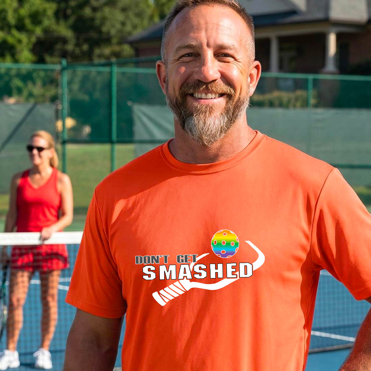 A smiling man wearing an orange t-shirt on a pickleball court. The t-shirt features a graphic with a colorful pickleball and the white text 'DON'T GET SMASHED' with a pickleball paddle icon. 
