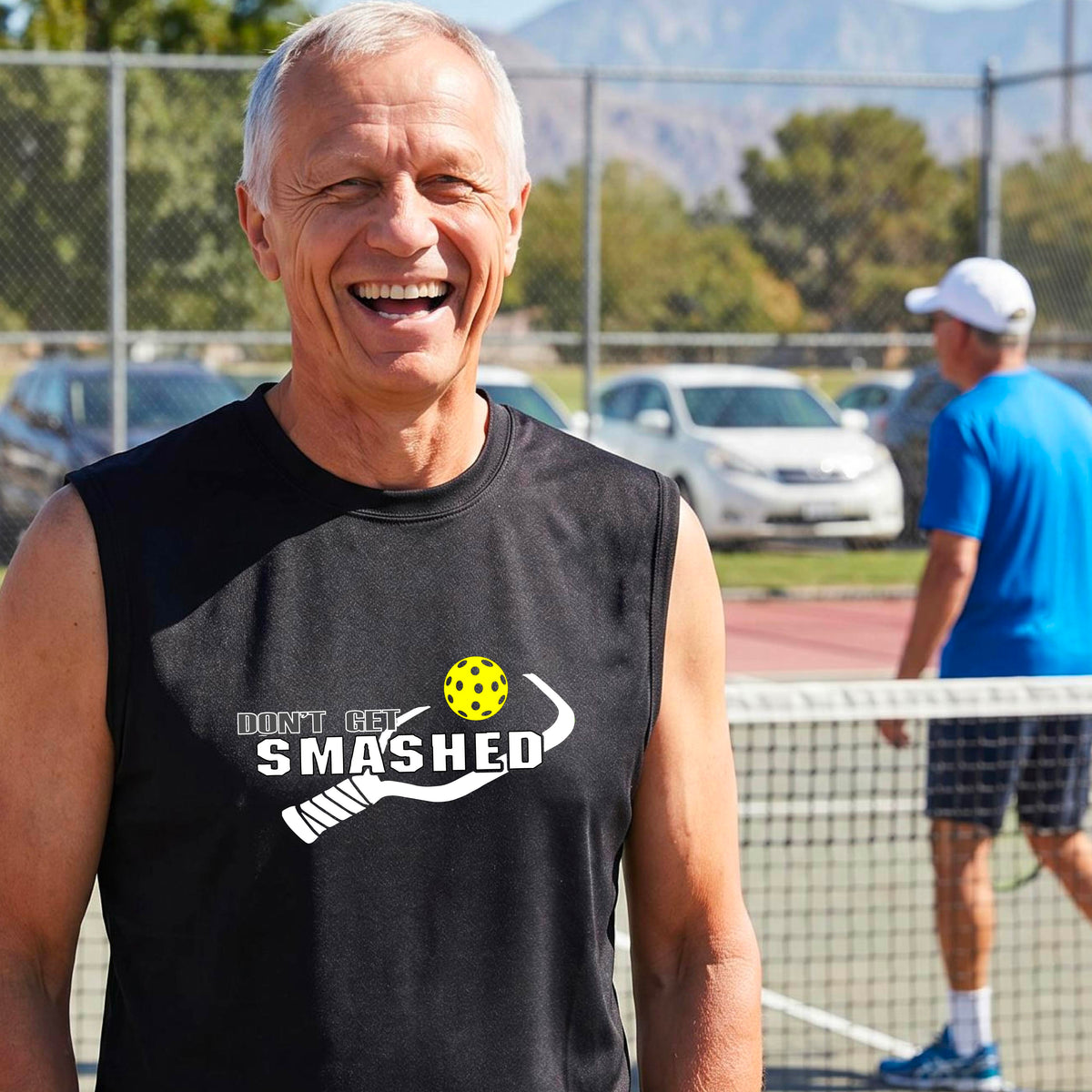A smiling man wearing a black sleeveless t-shirt with a yellow and white graphic that reads 'DON'T GET SMASHED' with a white pickleball paddle and colorful pickleball graphic.