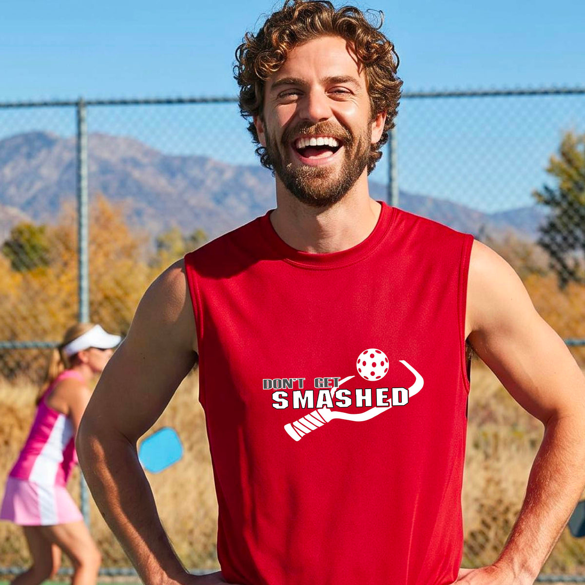 A smiling man wearing a red sleeveless t-shirt with a yellow and white graphic that reads 'DON'T GET SMASHED' with a white pickleball paddle and colorful pickleball graphic.