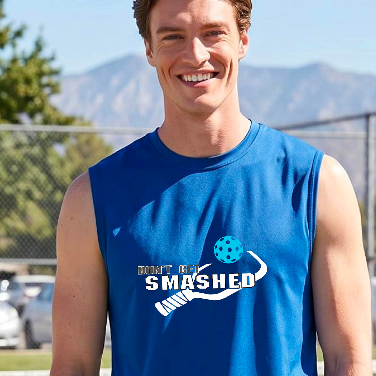 A smiling man wearing a royal blue sleeveless t-shirt with a yellow and white graphic that reads 'DON'T GET SMASHED' with a white pickleball paddle and colorful pickleball graphic.