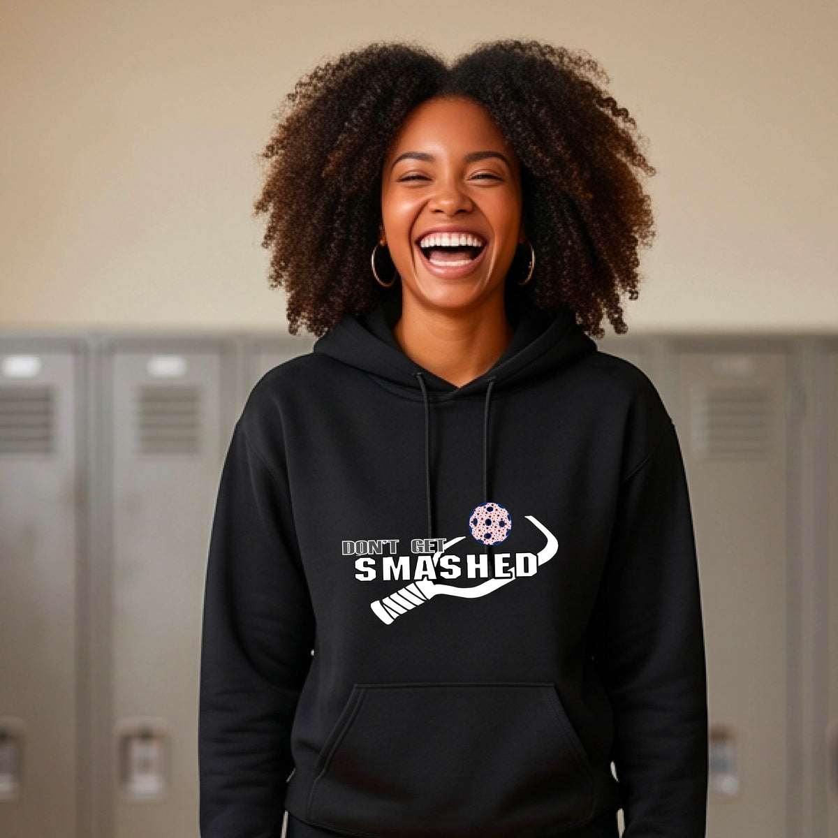 A woman wearing a black pullover hoodie with a white graphic print on the chest that reads 'DON'T GET SMASHED' with a pickleball paddle and colorful pickleball design.