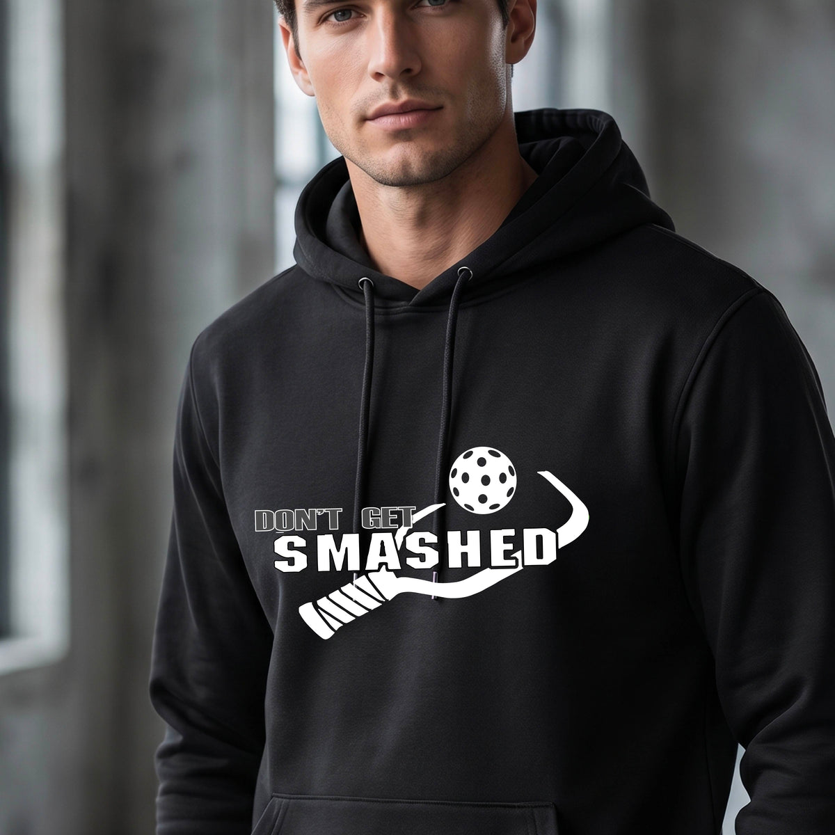 A man wearing a black pullover hoodie with a white graphic print on the chest that reads 'DON'T GET SMASHED' with a pickleball paddle and colorful pickleball design.