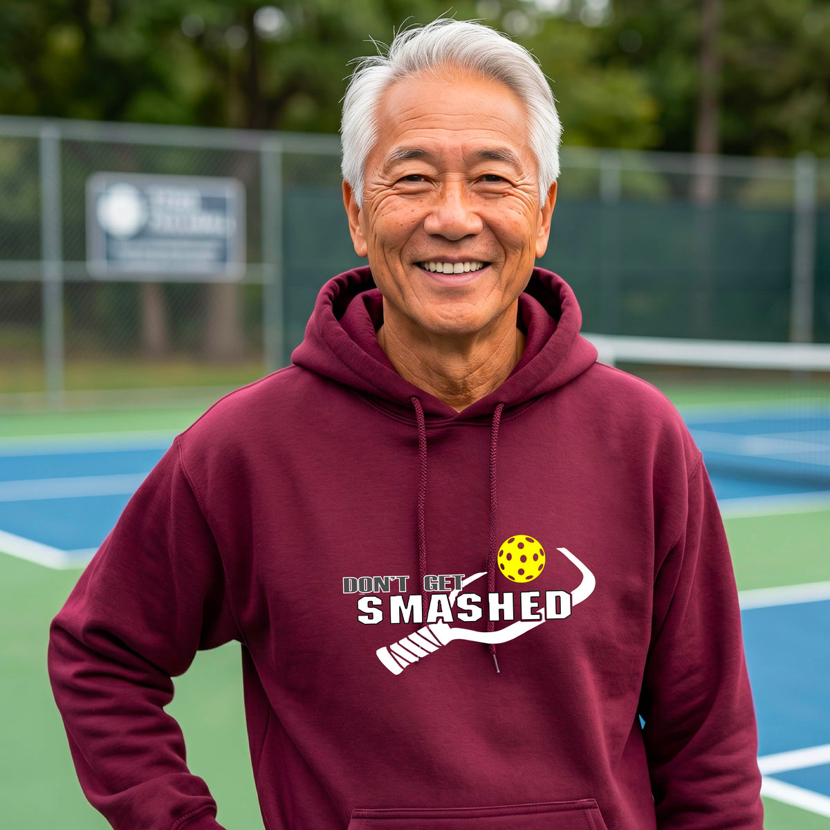 A man wearing a maroon pullover hoodie with a white graphic print on the chest that reads 'DON'T GET SMASHED' with a pickleball paddle and colorful pickleball design.
