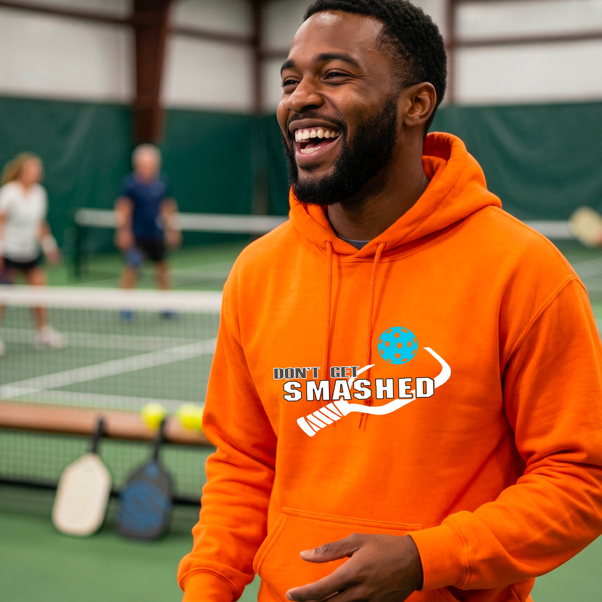 A man wearing an orange pullover hoodie with a white graphic print on the chest that reads 'DON'T GET SMASHED' with a pickleball paddle and colorful pickleball design.