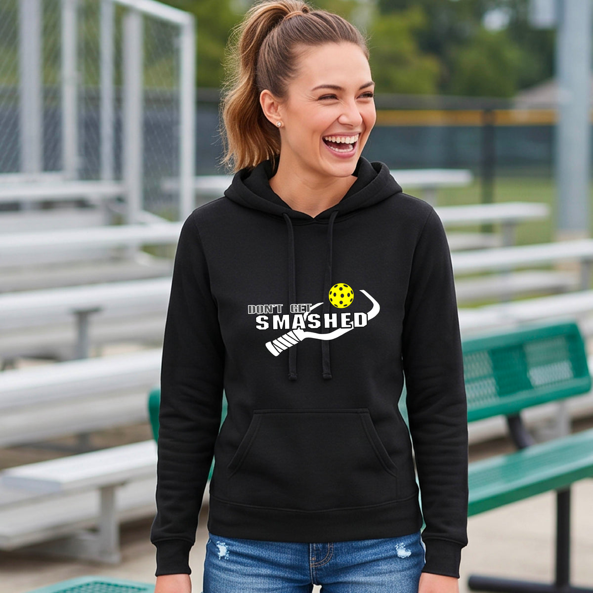 A smiling young woman wearing a black hoodie with a white and graphic that reads 'DON'T GET SMASHED' with white pickleball paddle and a colorful pickleball icon.