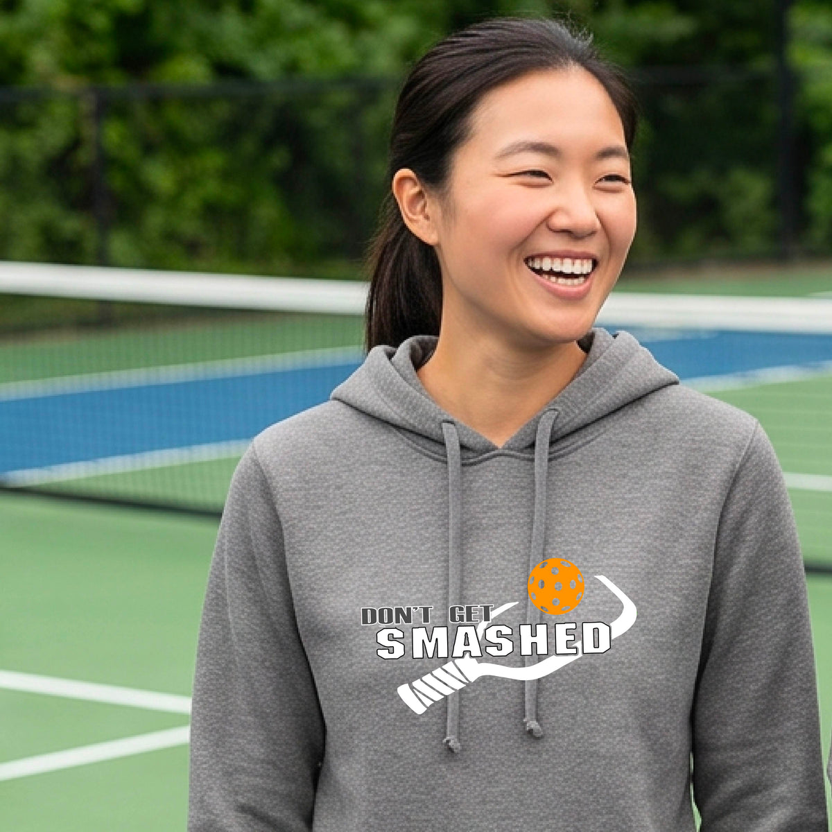 A smiling young woman wearing a gray hoodie with a white and graphic that reads 'DON'T GET SMASHED' with white pickleball paddle and a colorful pickleball icon.