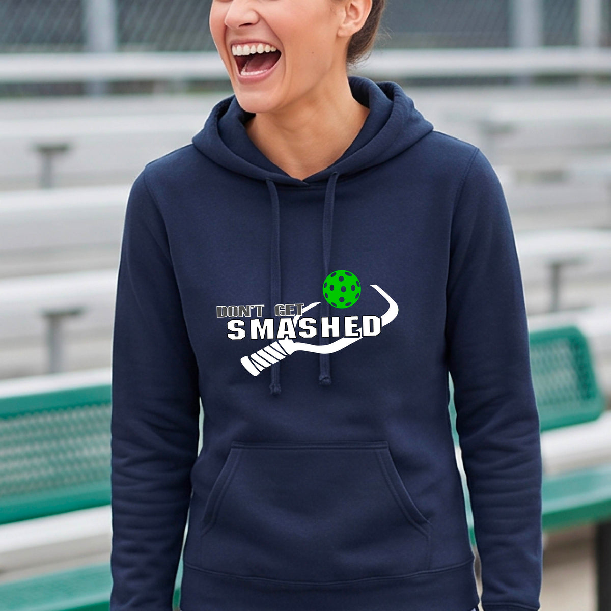 A smiling young woman wearing a navy blue hoodie with a white and graphic that reads 'DON'T GET SMASHED' with white pickleball paddle and a colorful pickleball icon.