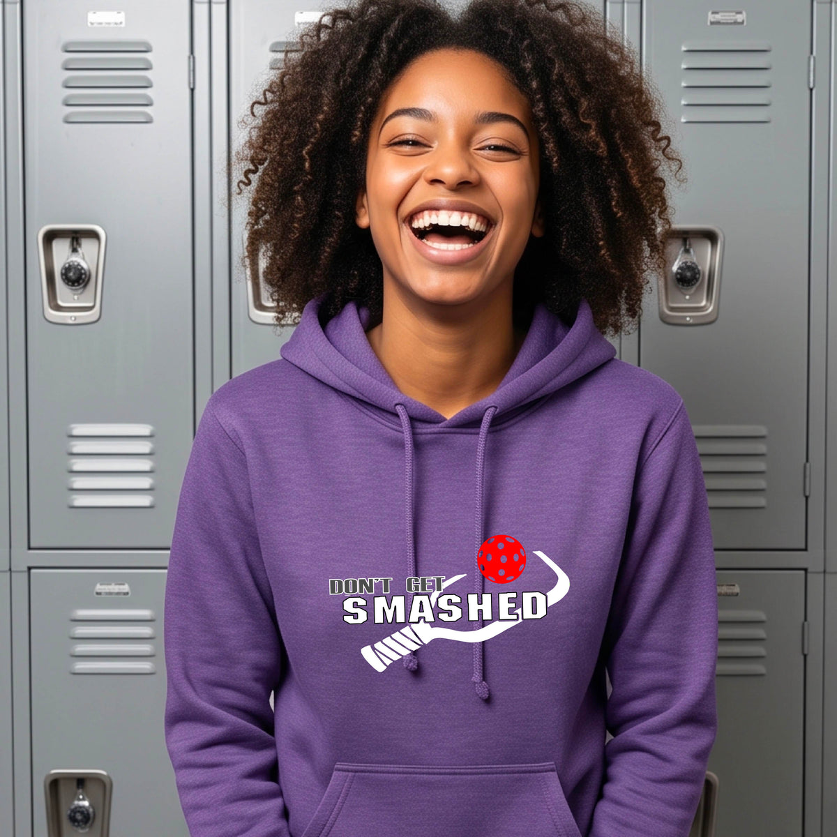 A smiling young woman wearing a purple hoodie with a white and graphic that reads 'DON'T GET SMASHED' with white pickleball paddle and a colorful pickleball icon.