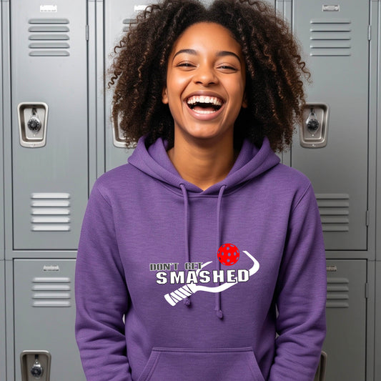 A smiling young woman wearing a purple hoodie with a white and graphic that reads 'DON'T GET SMASHED' with white pickleball paddle and a colorful pickleball icon.
