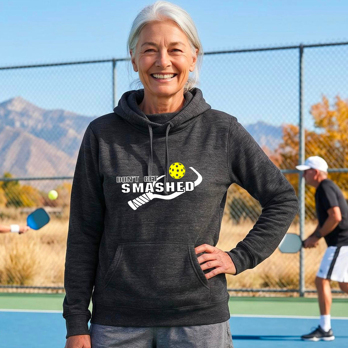 A woman laughing on a pickleball court wearing a heather black cowl-neck hoodie with a white' DON'T GET SMASHED' text with a white pickleball paddle and a colorful pickleball. 