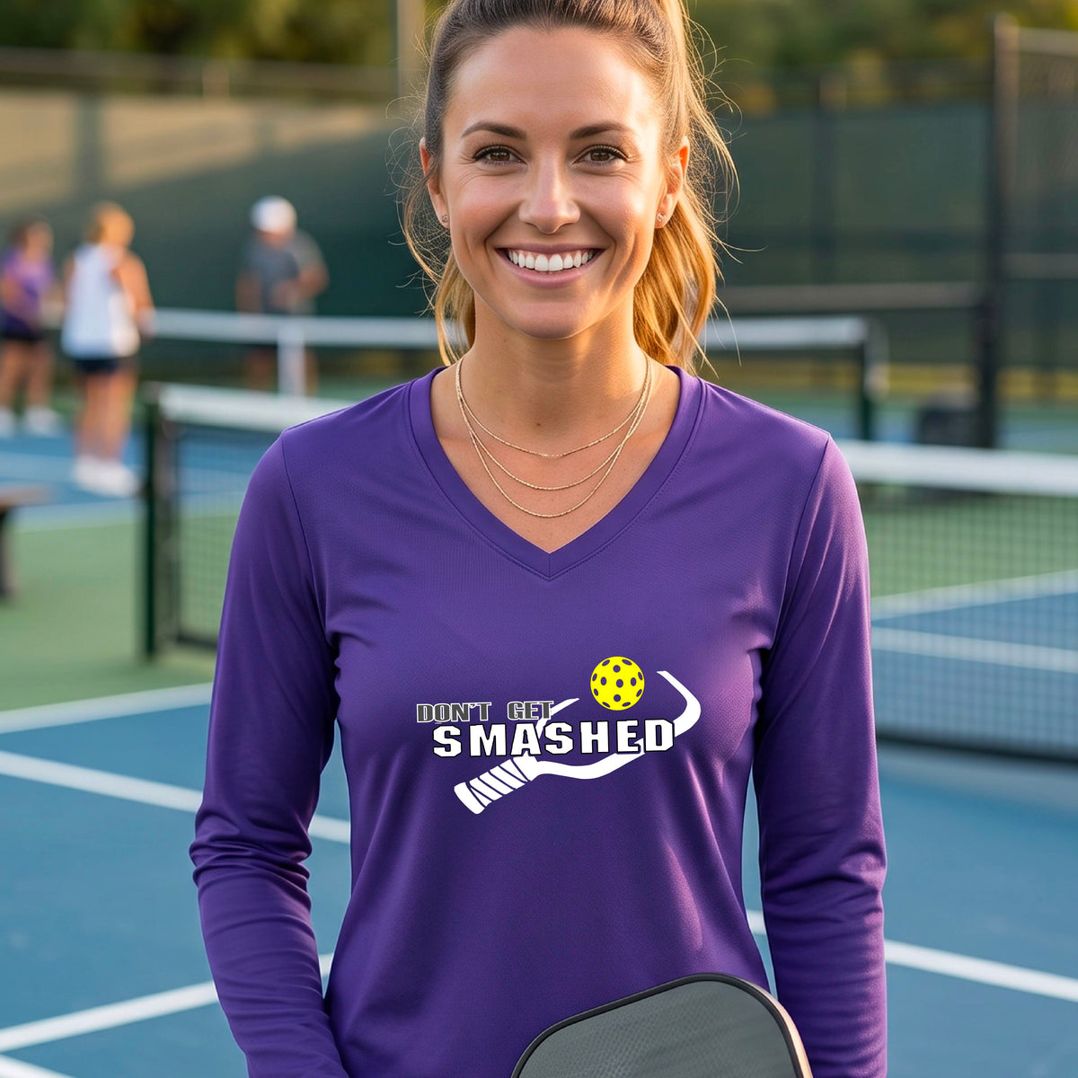A smiling woman wearing a purple long-sleeve V-neck athletic shirt with a white graphic that reads 'DON'T GET SMASHED' and a white pickleball paddle and colorful pickleball. 