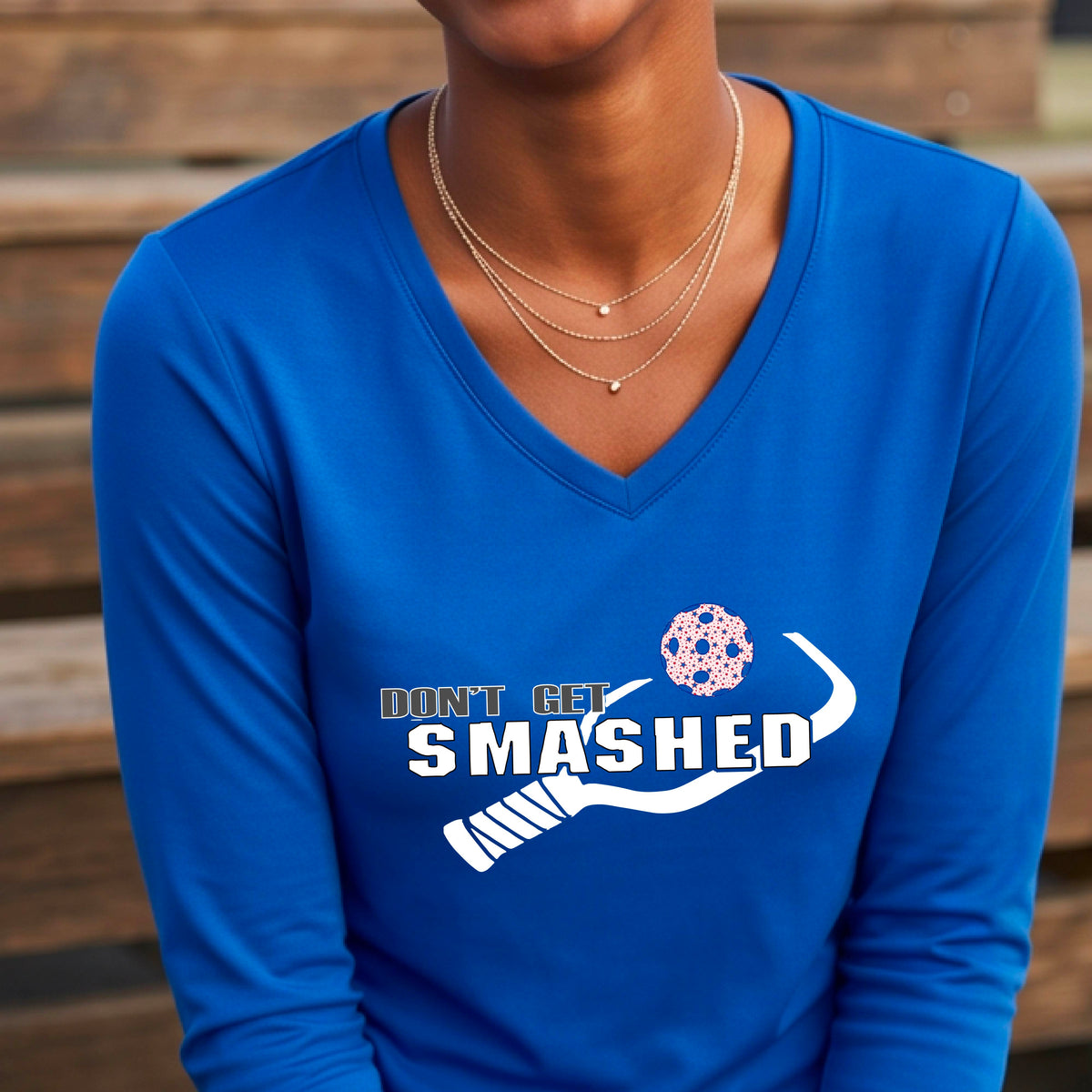 A smiling woman wearing a royal blue long-sleeve V-neck athletic shirt with a white graphic that reads 'DON'T GET SMASHED' and a white pickleball paddle and colorful pickleball. 