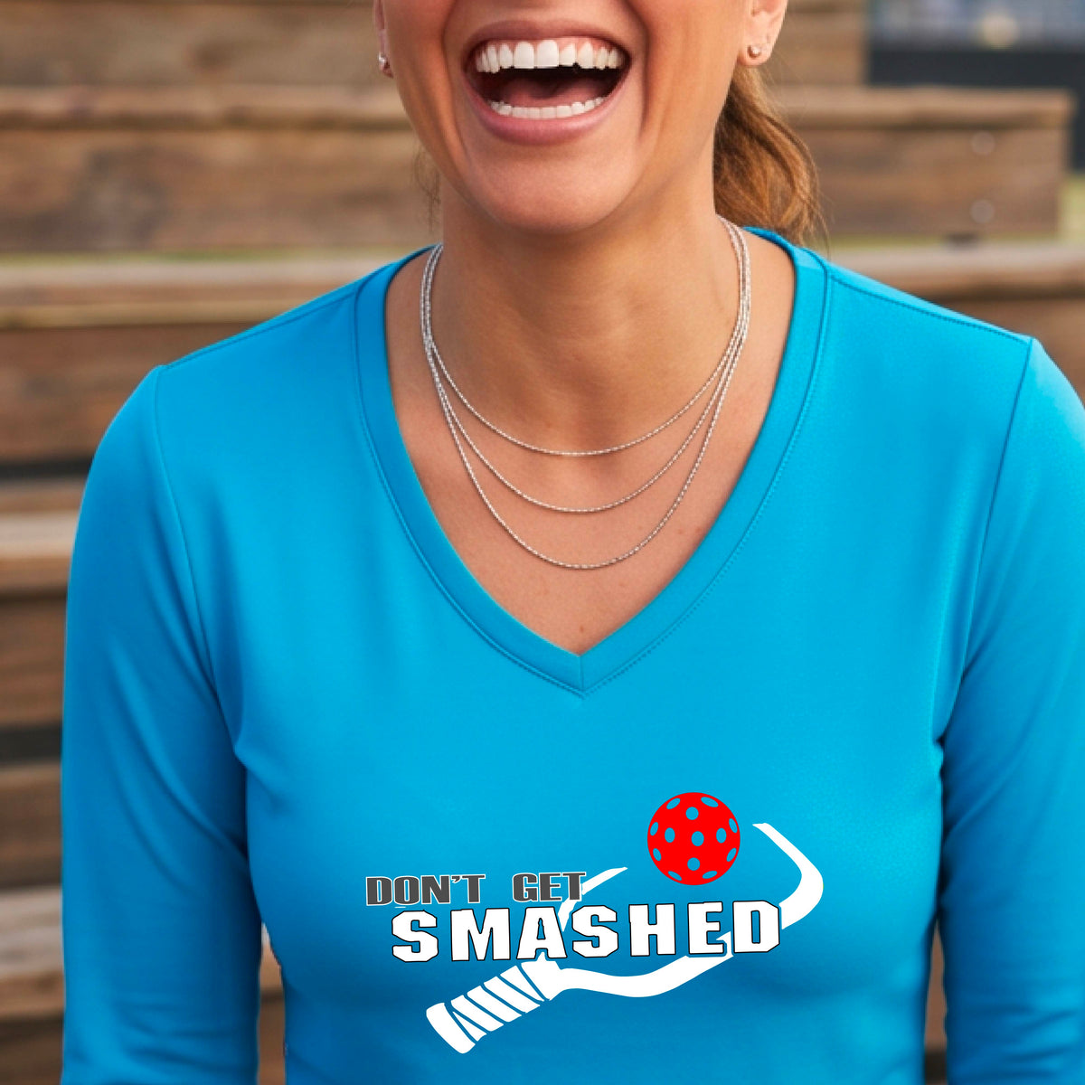 A smiling woman wearing a bright blue long-sleeve V-neck athletic shirt with a white graphic that reads 'DON'T GET SMASHED' and a white pickleball paddle and colorful pickleball. 