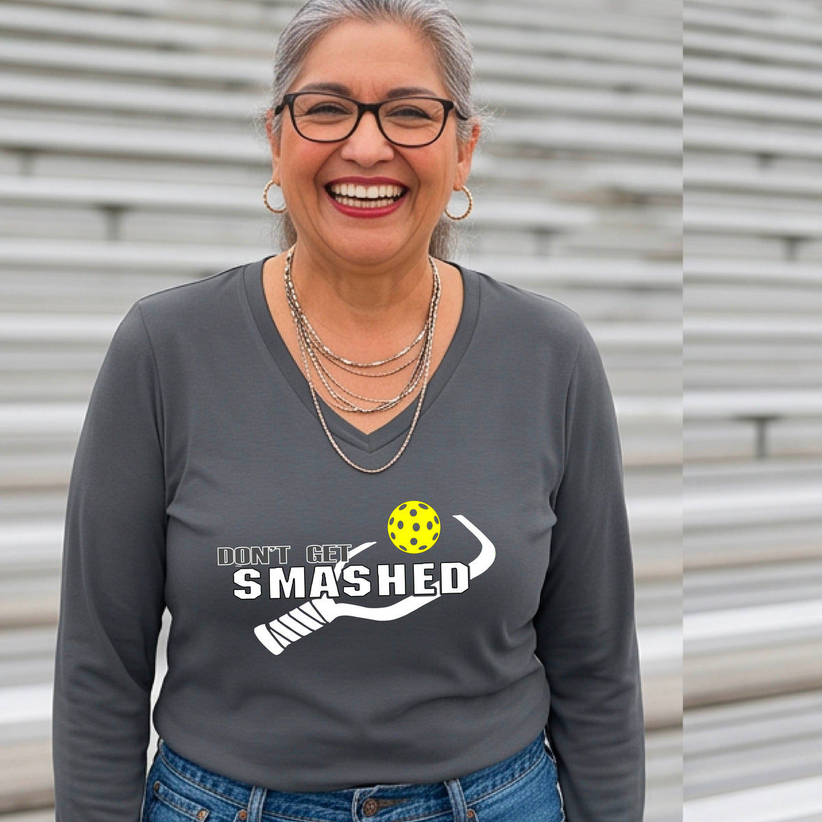 A smiling woman wearing a dark gray long-sleeve V-neck athletic shirt with a white graphic that reads 'DON'T GET SMASHED' and a white pickleball paddle and colorful pickleball. 