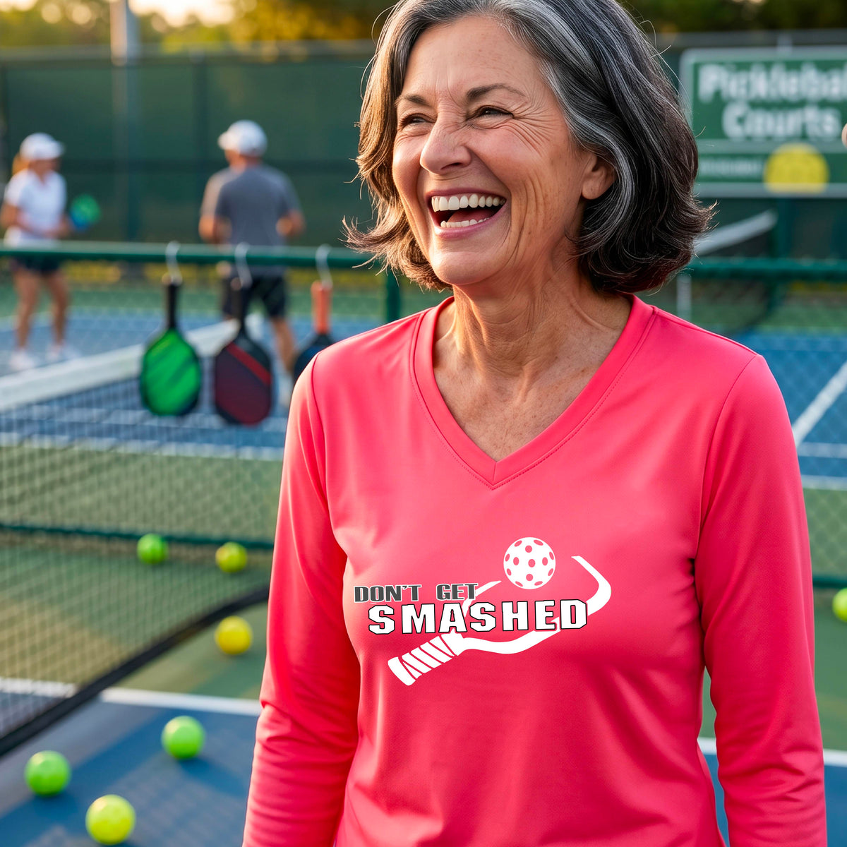 A smiling woman wearing a coral pink long-sleeve V-neck athletic shirt with a white graphic that reads 'DON'T GET SMASHED' and a white pickleball paddle and colorful pickleball. 
