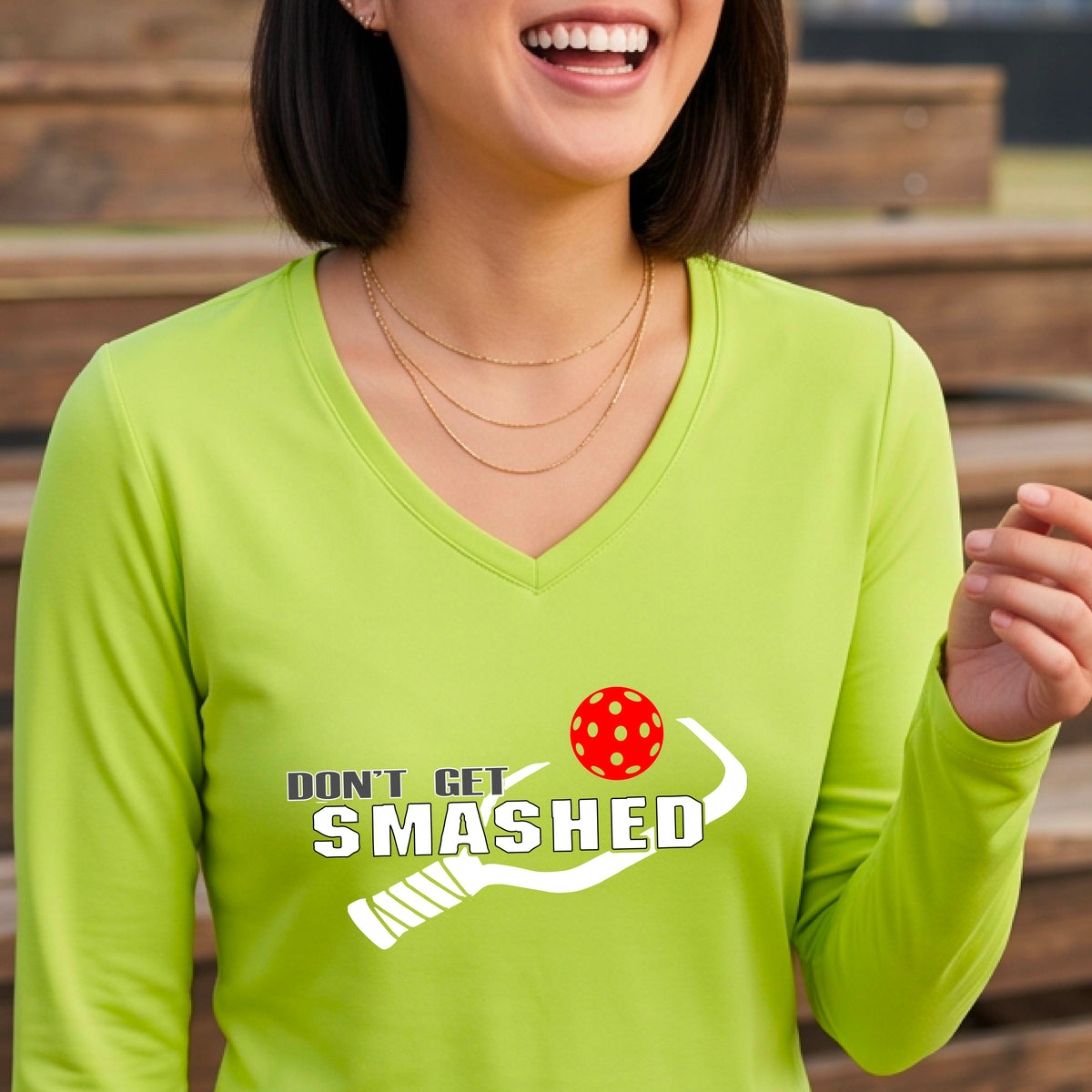 A smiling woman wearing a lime green long-sleeve V-neck athletic shirt with a white graphic that reads 'DON'T GET SMASHED' and a white pickleball paddle and colorful pickleball. 