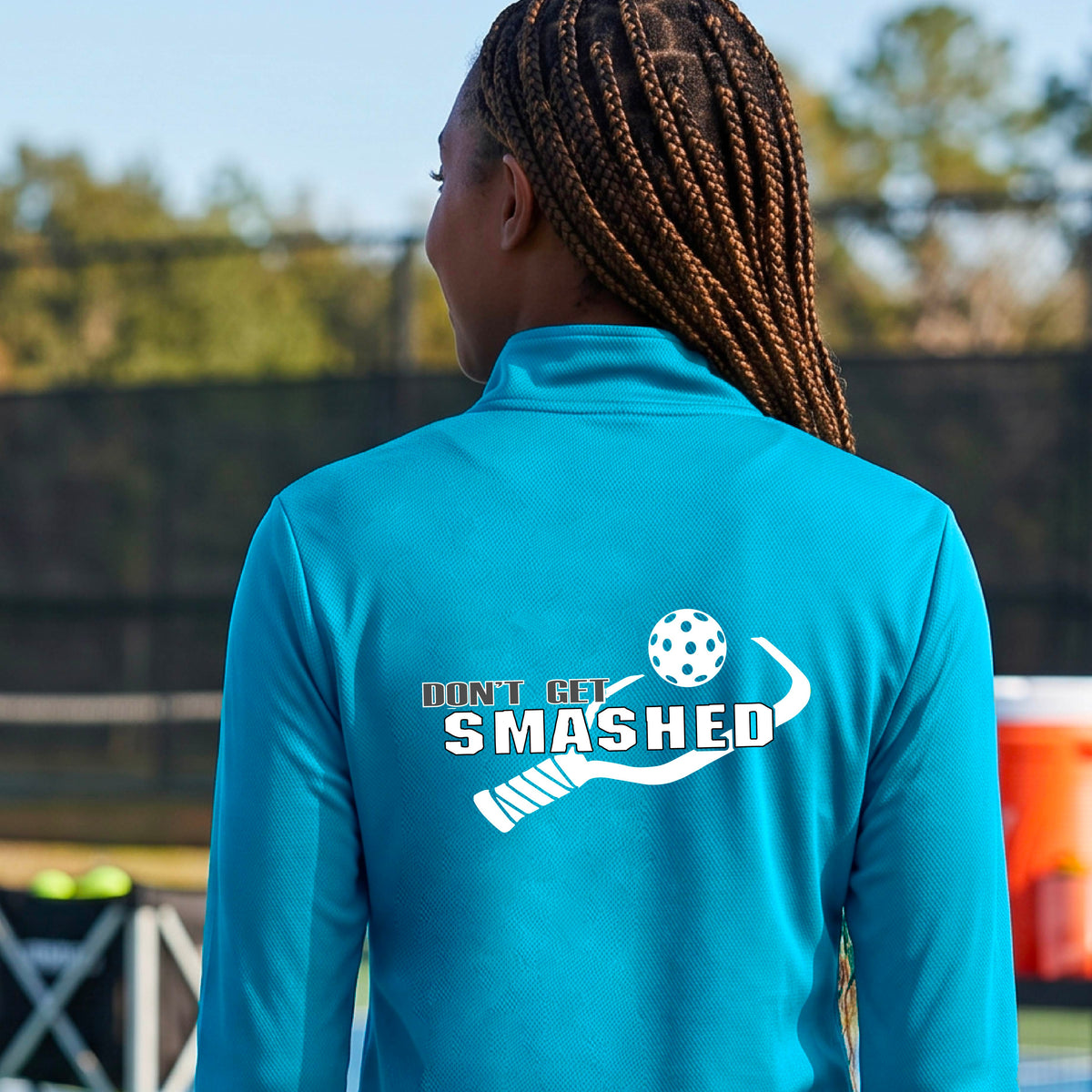 A woman viewed from behind wearing a bright turquoise athletic quarter-zip jacket with a white graphic on the back that reads 'DON'T GET SMASHED' with a pickleball paddle and colorful pickleball graphic.