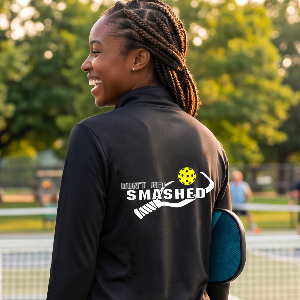 A woman viewed from behind wearing a black athletic quarter-zip jacket with a white graphic on the back that reads 'DON'T GET SMASHED' with a pickleball paddle and colorful pickleball graphic.