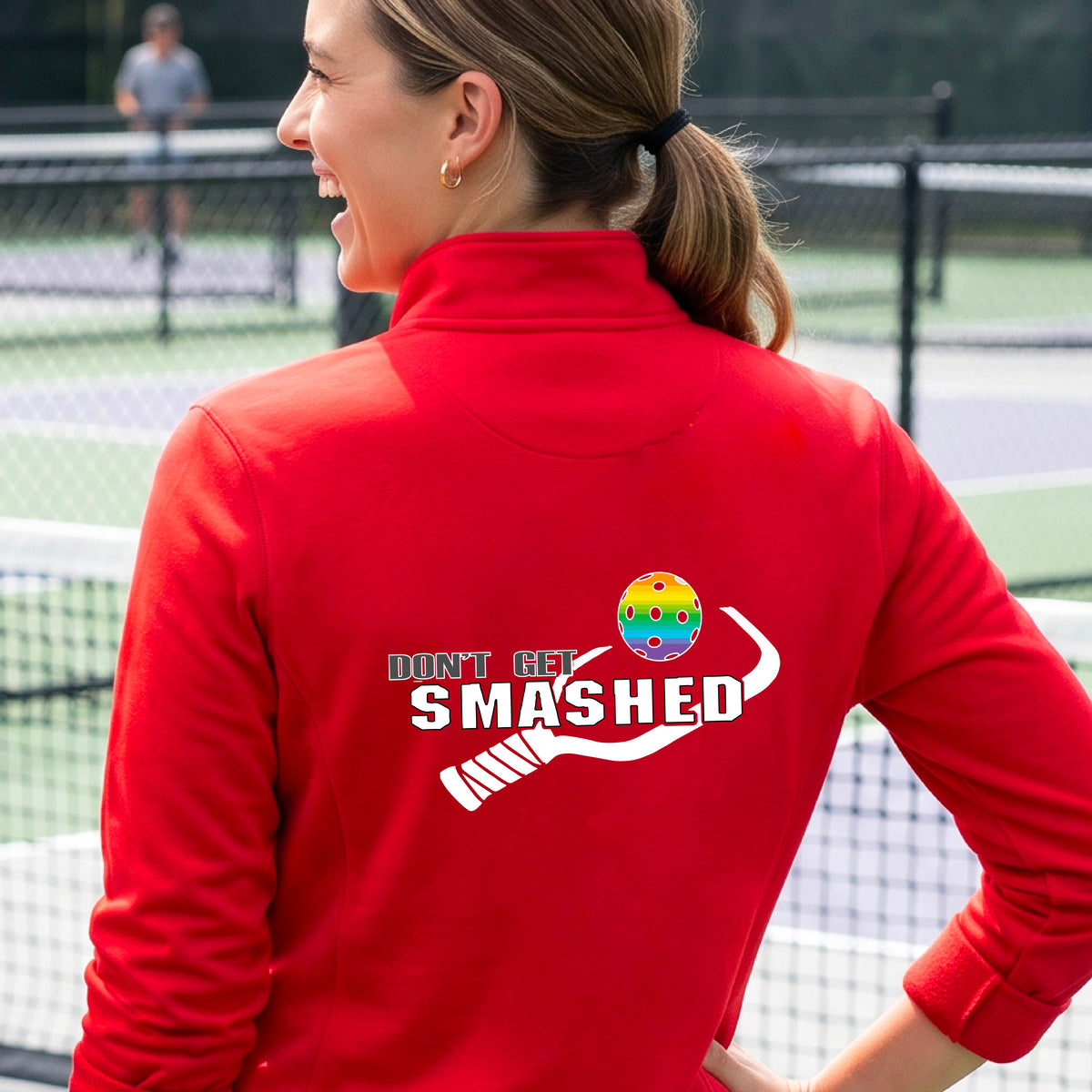 A woman viewed from behind wearing a bright red athletic quarter-zip jacket with a white graphic on the back that reads 'DON'T GET SMASHED' with a pickleball paddle and colorful pickleball graphic.