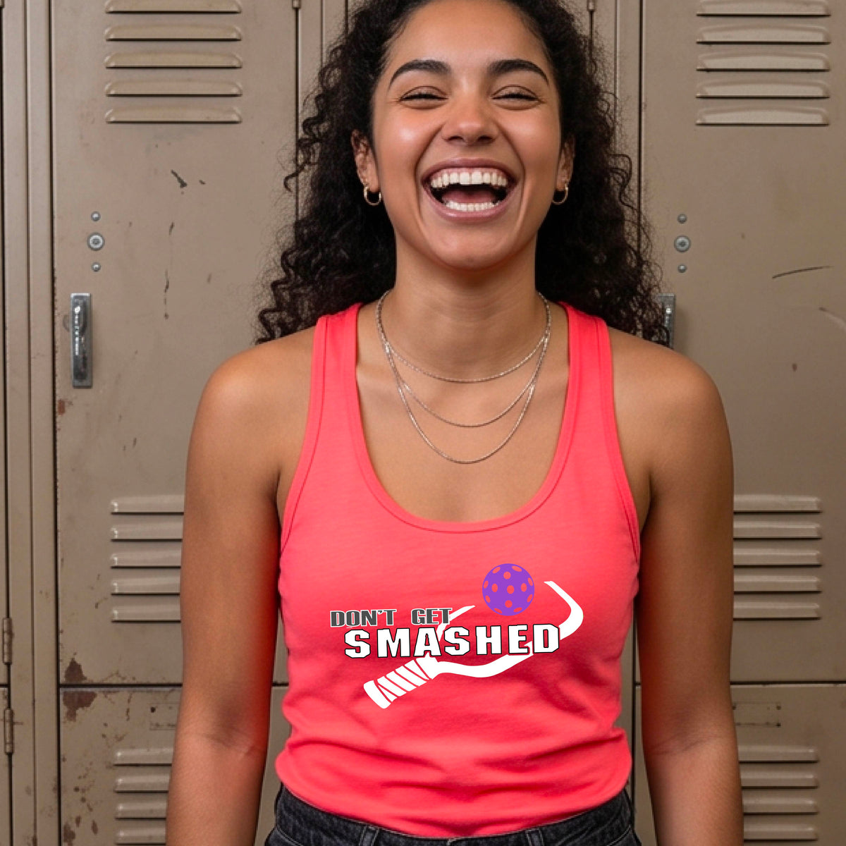 A woman on a pickleball court wearing a bright coral pink athletic racerback tank top with a white and green 'DON'T GET SMASHED' graphic with a pickleball paddle and colorful pickleball. 