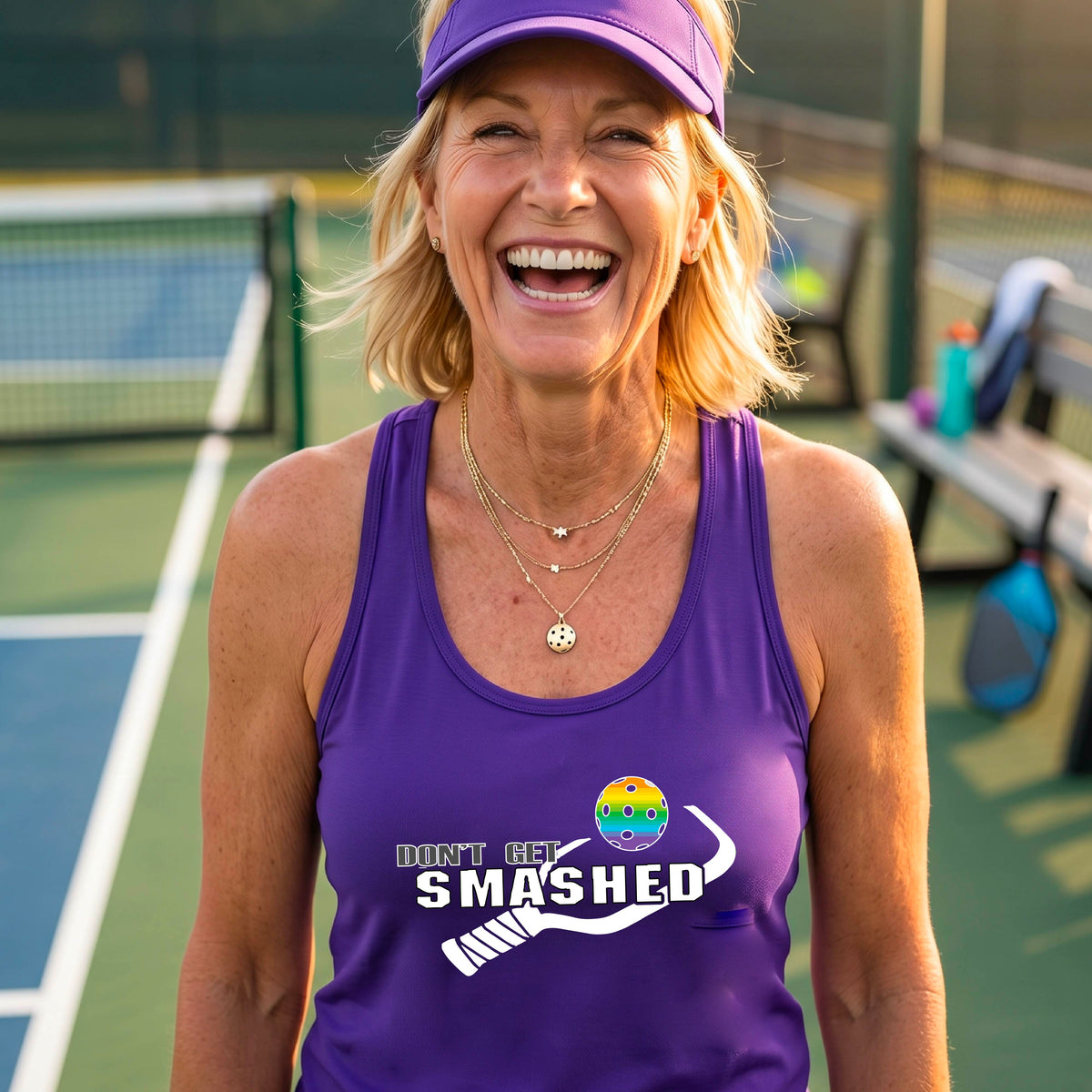 A woman on a pickleball court wearing a purple athletic racerback tank top with a white and green 'DON'T GET SMASHED' graphic with a pickleball paddle and colorful pickleball. 