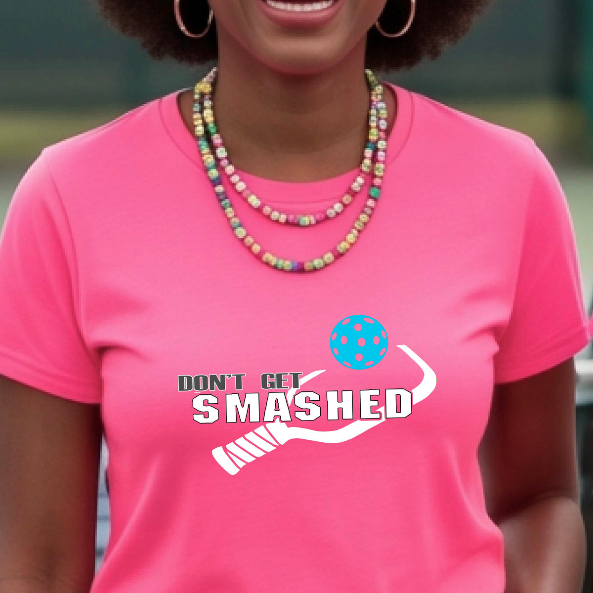 A close-up of a woman wearing a bright pink t-shirt with a graphic that reads 'DON'T GET SMASHED' in white accompanied by a colorful pickleball and a white pickleball paddle.