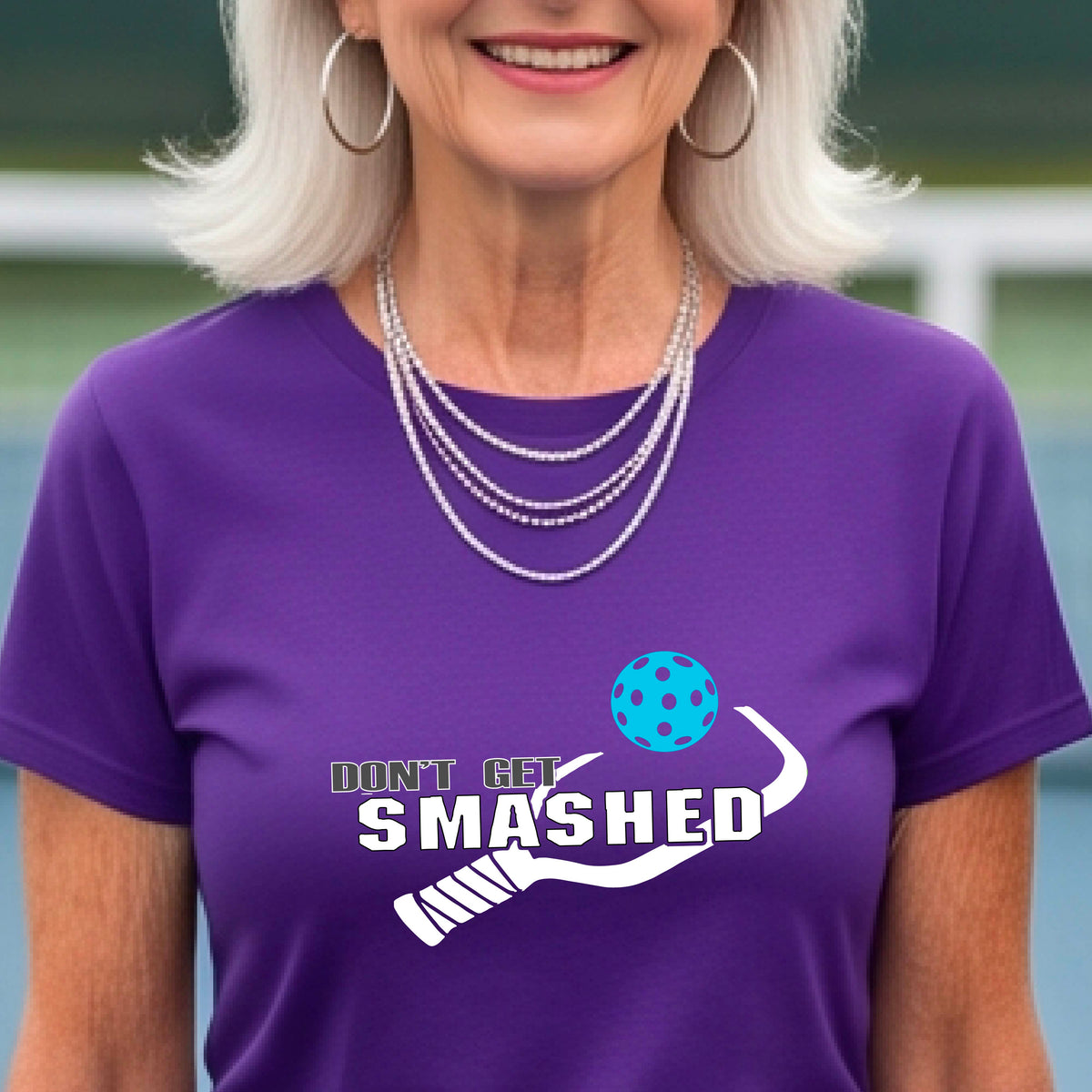 A close-up of a woman wearing a purple t-shirt with a graphic that reads 'DON'T GET SMASHED' in white accompanied by a colorful pickleball and a white pickleball paddle.