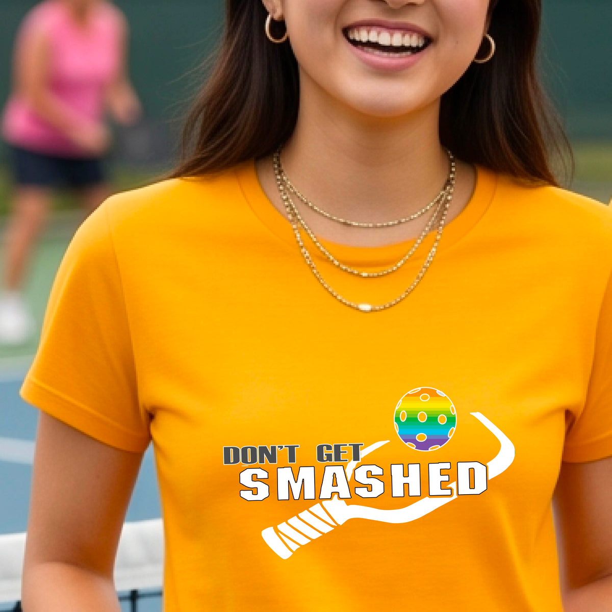 A close-up of a woman wearing a mustard yellow gold t-shirt with a graphic that reads 'DON'T GET SMASHED' in white accompanied by a colorful pickleball and a white pickleball paddle.