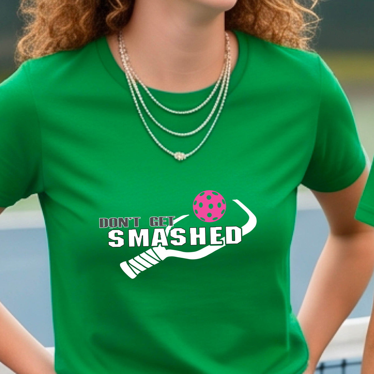 A close-up of a woman wearing a green t-shirt with a graphic that reads 'DON'T GET SMASHED' in white accompanied by a colorful pickleball and a white pickleball paddle.