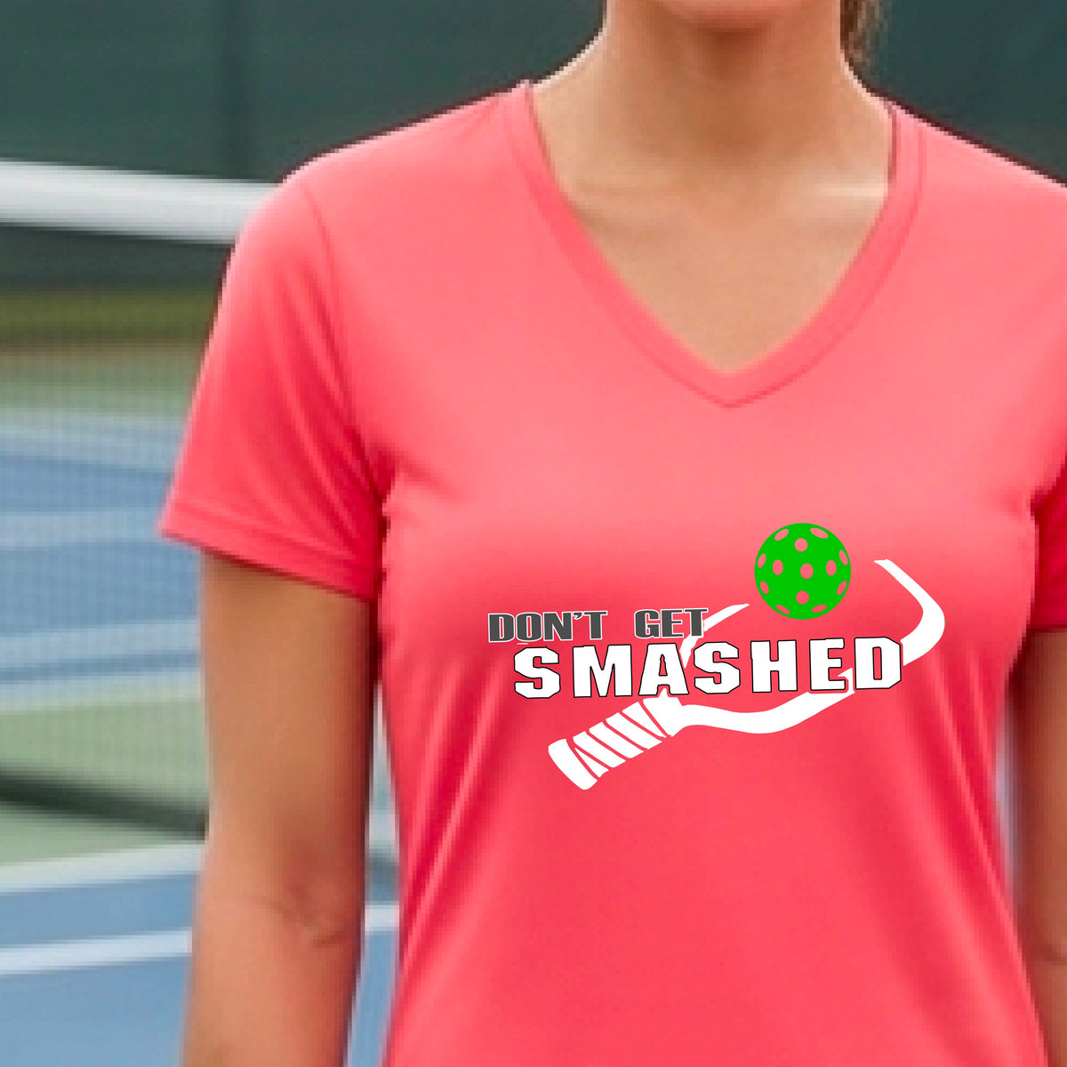 A woman wearing a coral pink V-neck t-shirt with a white graphic that reads 'DON'T GET SMASHED' alongside a pickleball paddle and a colorful pickleball.
