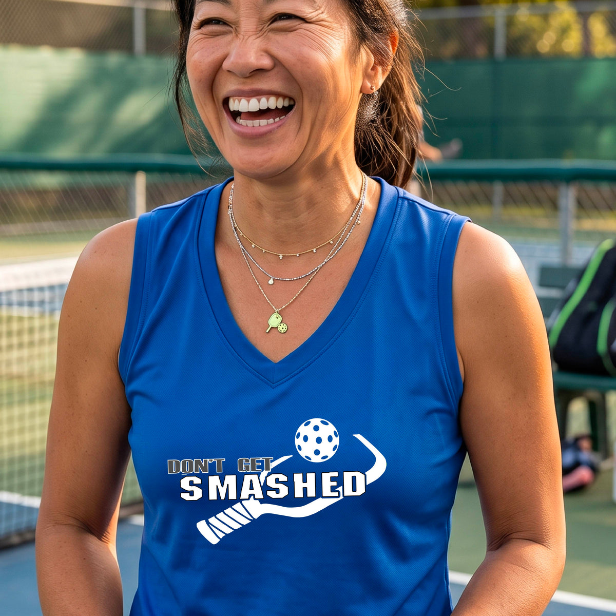 A smiling woman wearing a royal blue sleeveless V-neck athletic tank top with a pickleball -themed graphic that reads 'DON'T GET SMASHED' and shows a white pickleball paddle and colorful pickleball. 