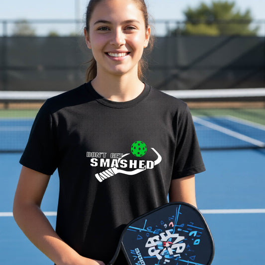A smiling young person on a blue pickleball court wearing a black short-sleeve t-shirt with a white and green 'DON'T GET SMASHED' with a pickleball paddle and colorful pickleball graphic. 