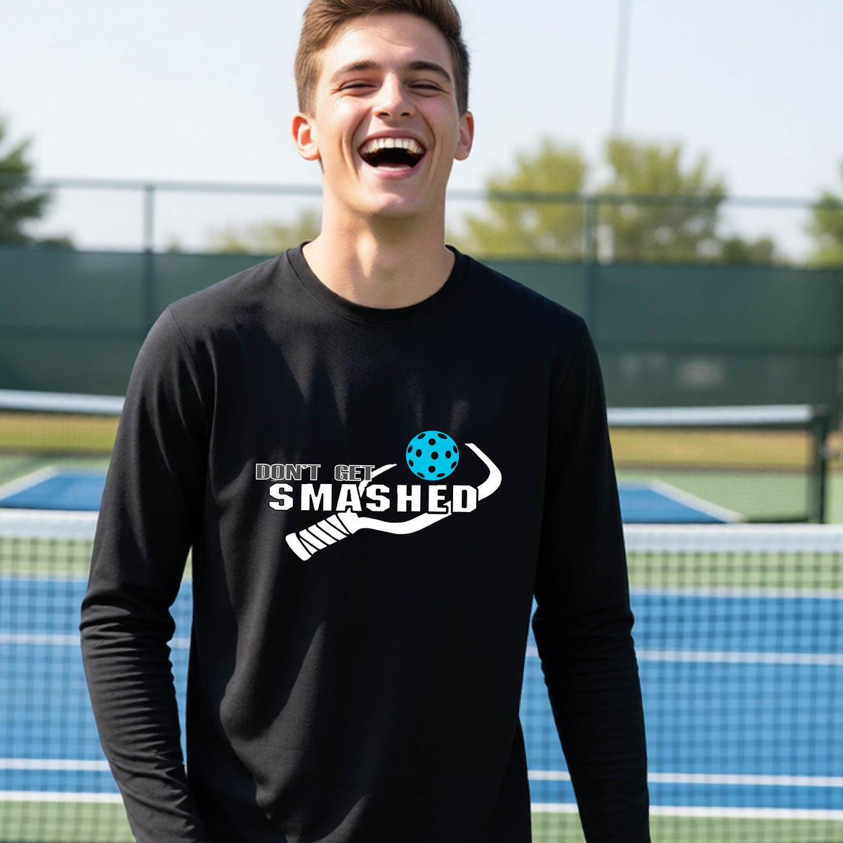 A young person on an outdoor pickleball court wearing a black long-sleeve athletic shirt with a white 'DONT GET SMASHED' graphic including a pickleball paddle and colorful pickleball on the chest.