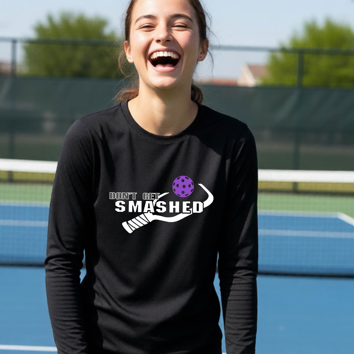 A young person on an outdoor pickleball court wearing a black long-sleeve athletic shirt with a white 'DONT GET SMASHED' graphic including a pickleball paddle and colorful pickleball on the chest.