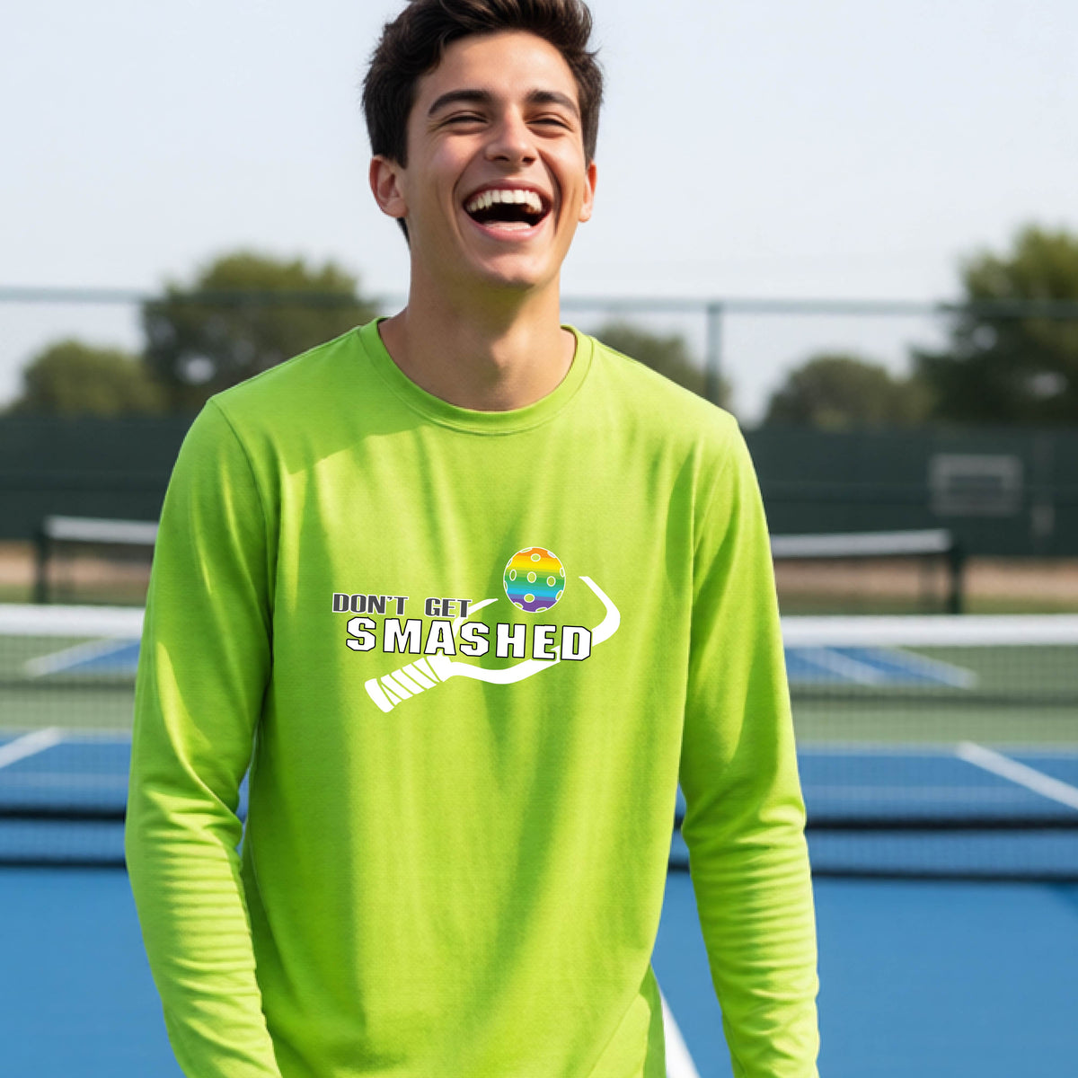 A young person on an outdoor pickleball court wearing a lime green long-sleeve athletic shirt with a white 'DONT GET SMASHED' graphic including a pickleball paddle and colorful pickleball on the chest.