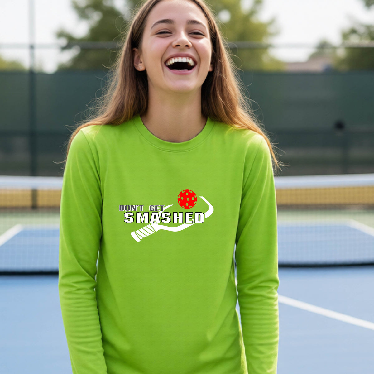 A young person on an outdoor pickleball court wearing a lime green long-sleeve athletic shirt with a white 'DONT GET SMASHED' graphic including a pickleball paddle and colorful pickleball on the chest.