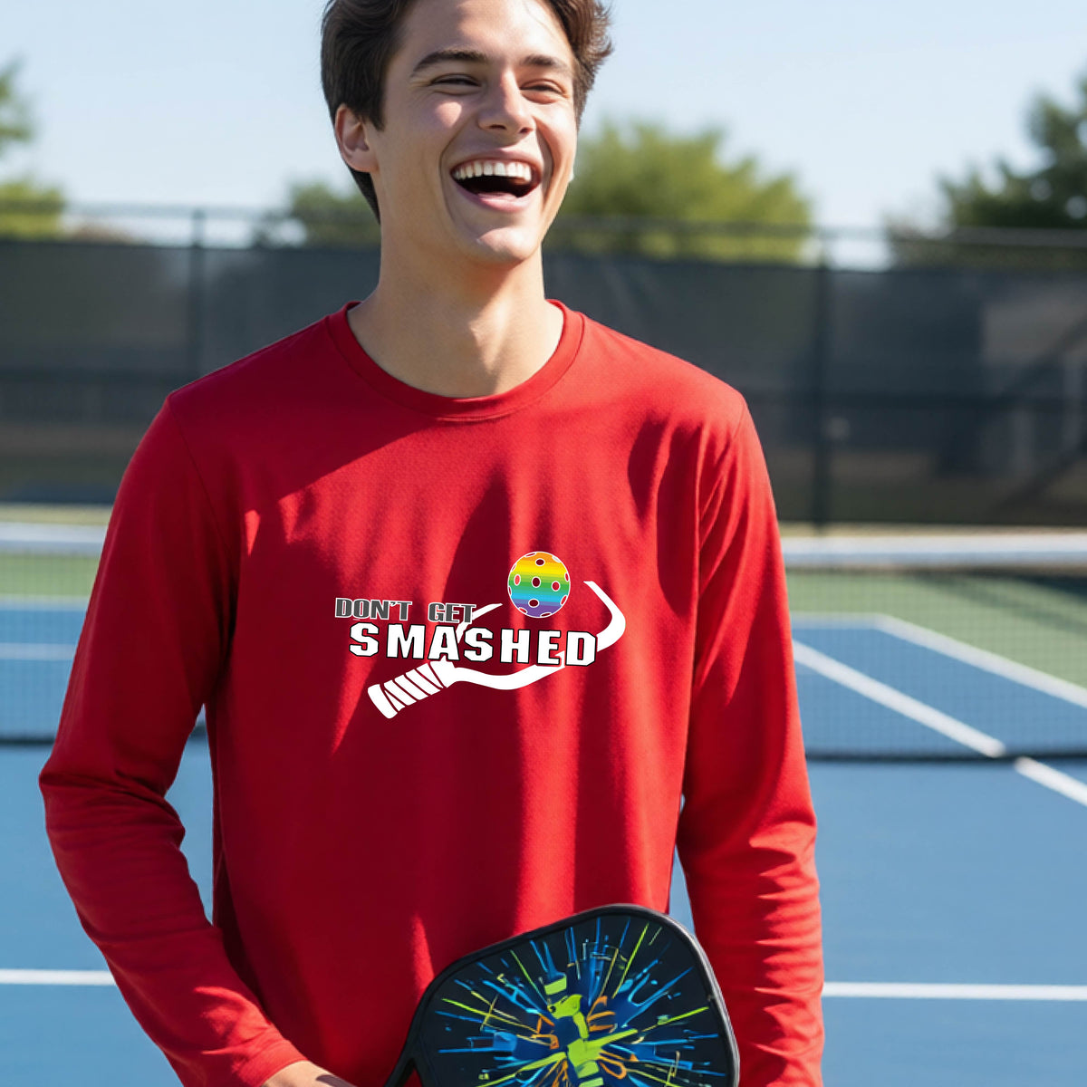 A young person on an outdoor pickleball court wearing a red long-sleeve athletic shirt with a white 'DONT GET SMASHED' graphic including a pickleball paddle and colorful pickleball on the chest.