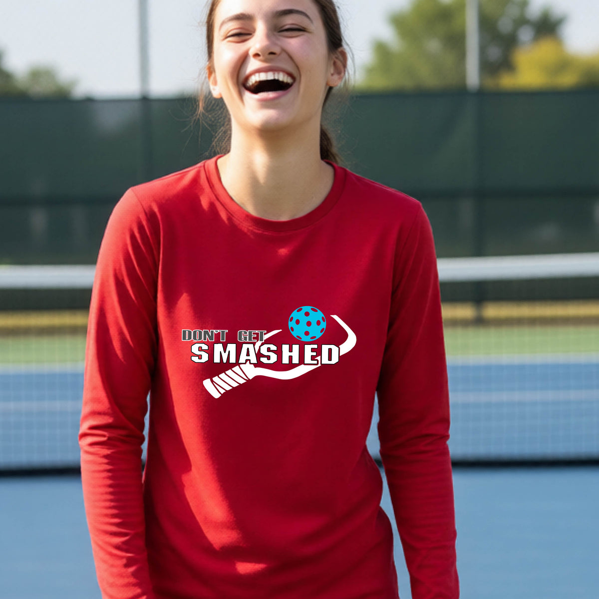 A young person on an outdoor pickleball court wearing a red long-sleeve athletic shirt with a white 'DONT GET SMASHED' graphic including a pickleball paddle and colorful pickleball on the chest.