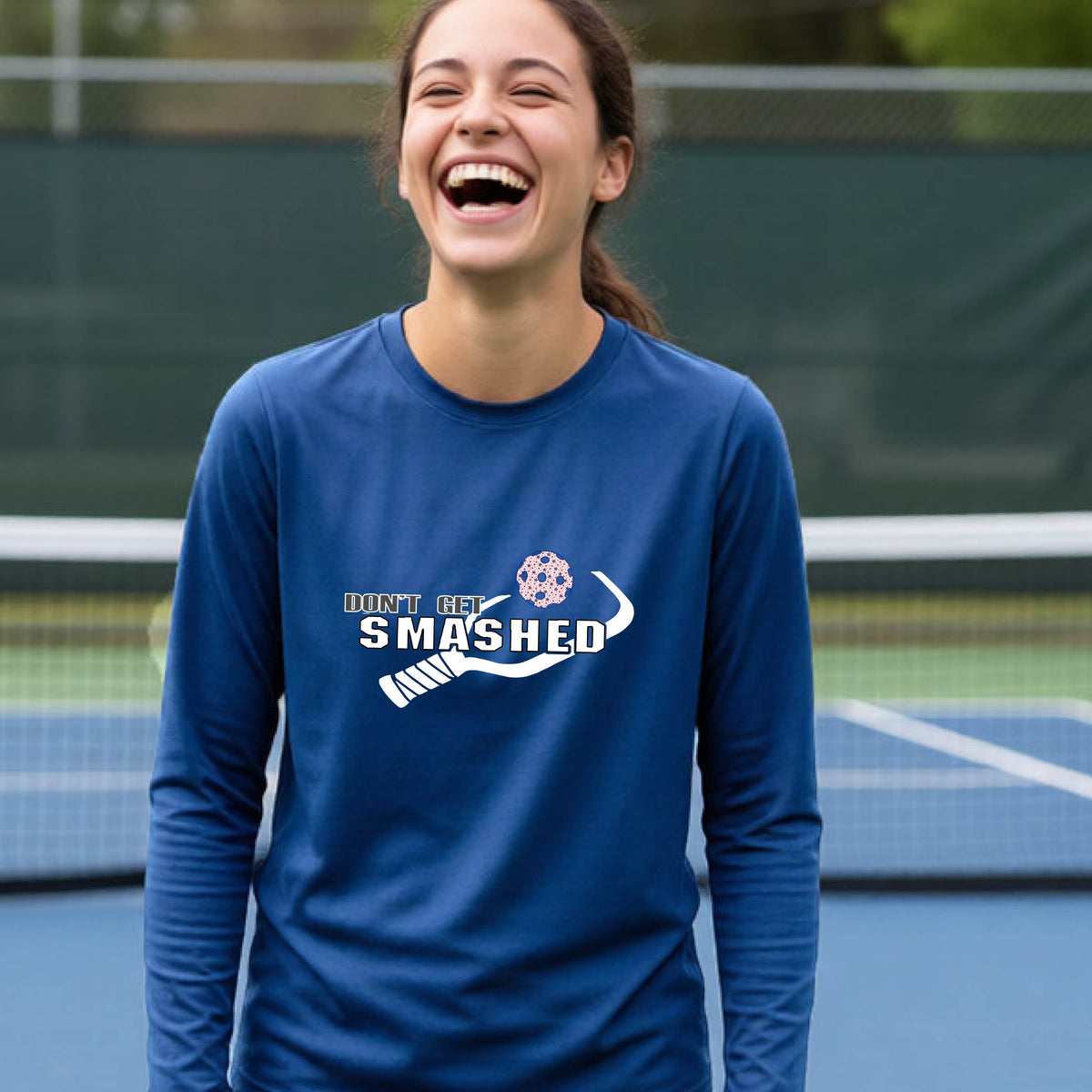 A young person on an outdoor pickleball court wearing a royal blue long-sleeve athletic shirt with a white 'DONT GET SMASHED' graphic including a pickleball paddle and colorful pickleball on the chest.